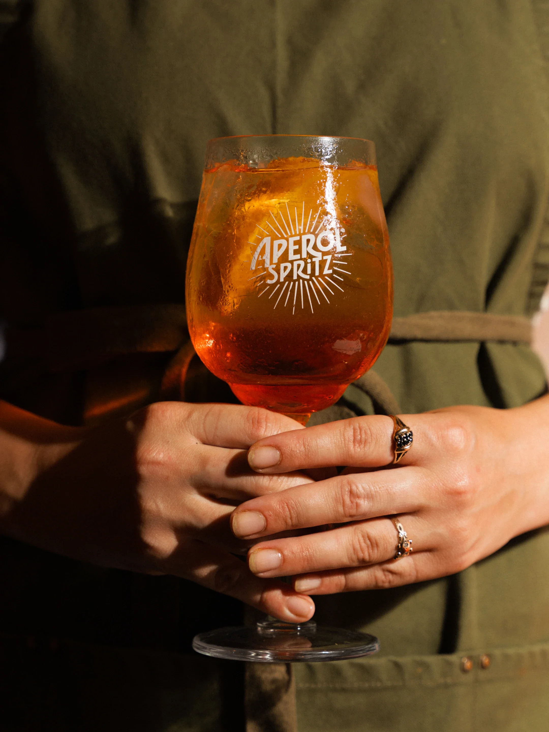 A hand holds a stemmed glass of orange Aperol Spritz cocktail with ice.