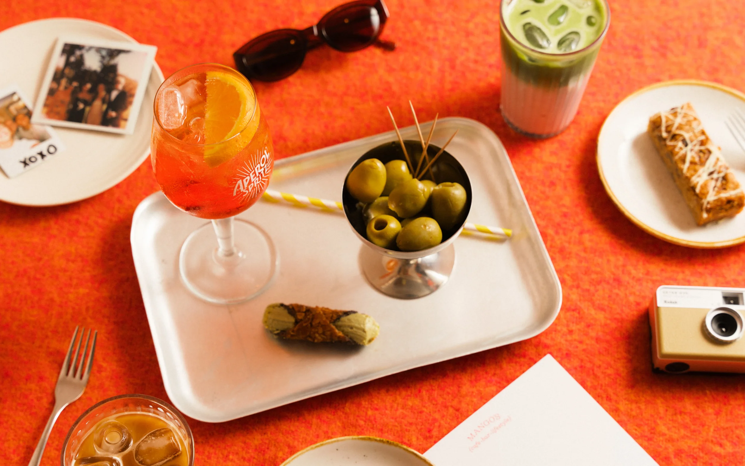 Overhead view of cocktails, green olives, pastries, sunglasses, and a vintage camera on a bright red table