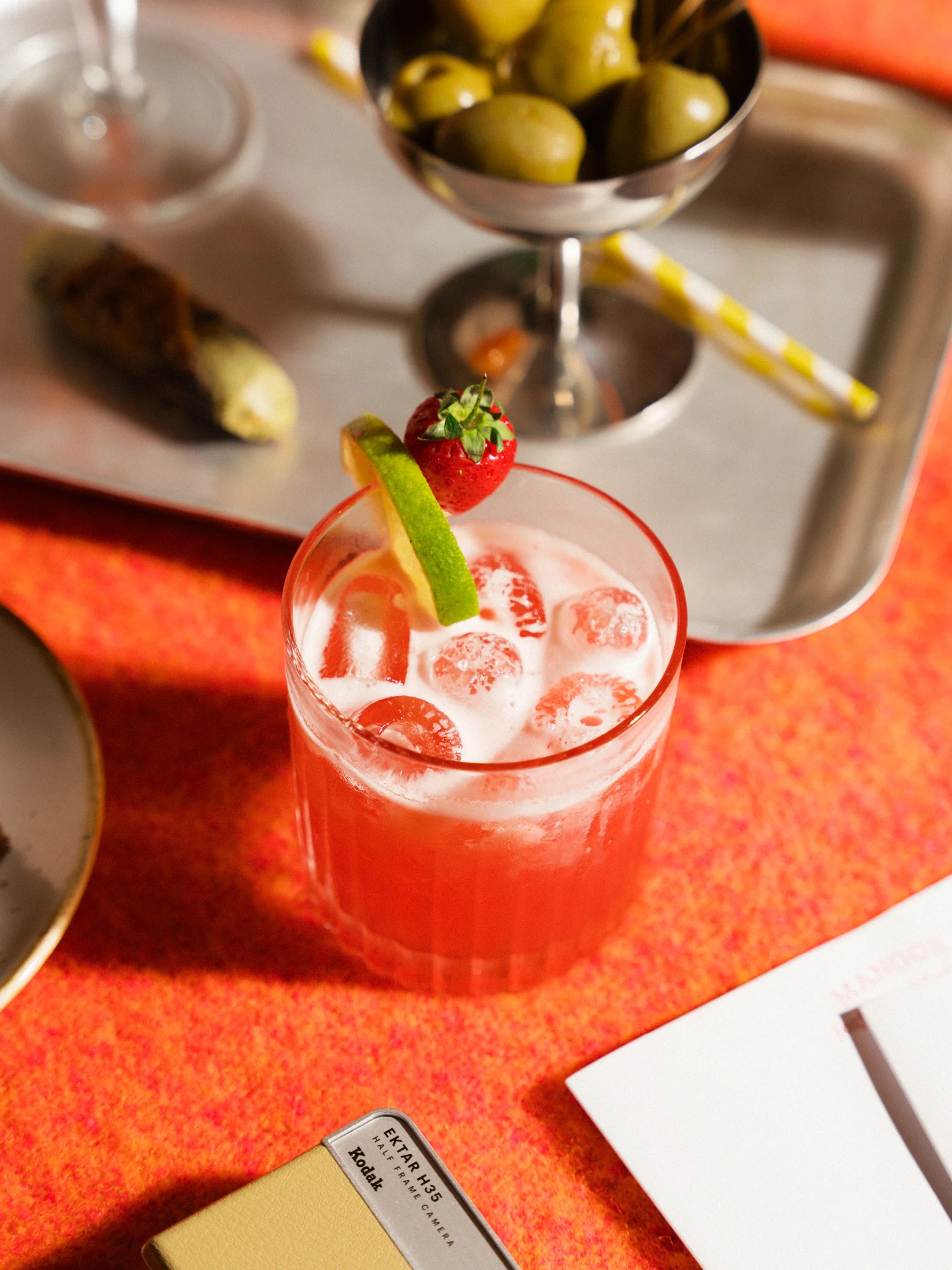 Red cocktail with ice, lime wedge, and strawberry garnish beside a bowl of green olives