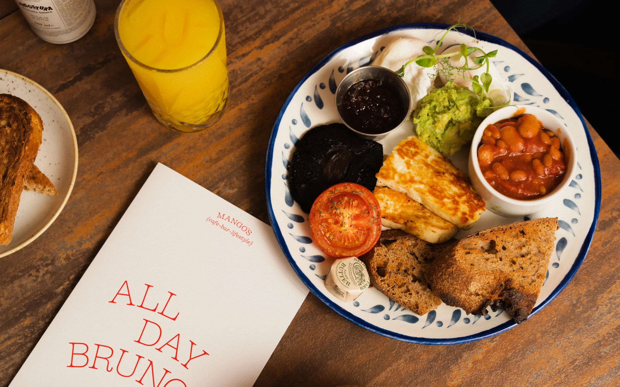 Vegetarian brunch platter with grilled halloumi, avocado, baked beans, tomato, sourdough toast, black pudding, and microgreens, served with orange juice and coffee-table menus.