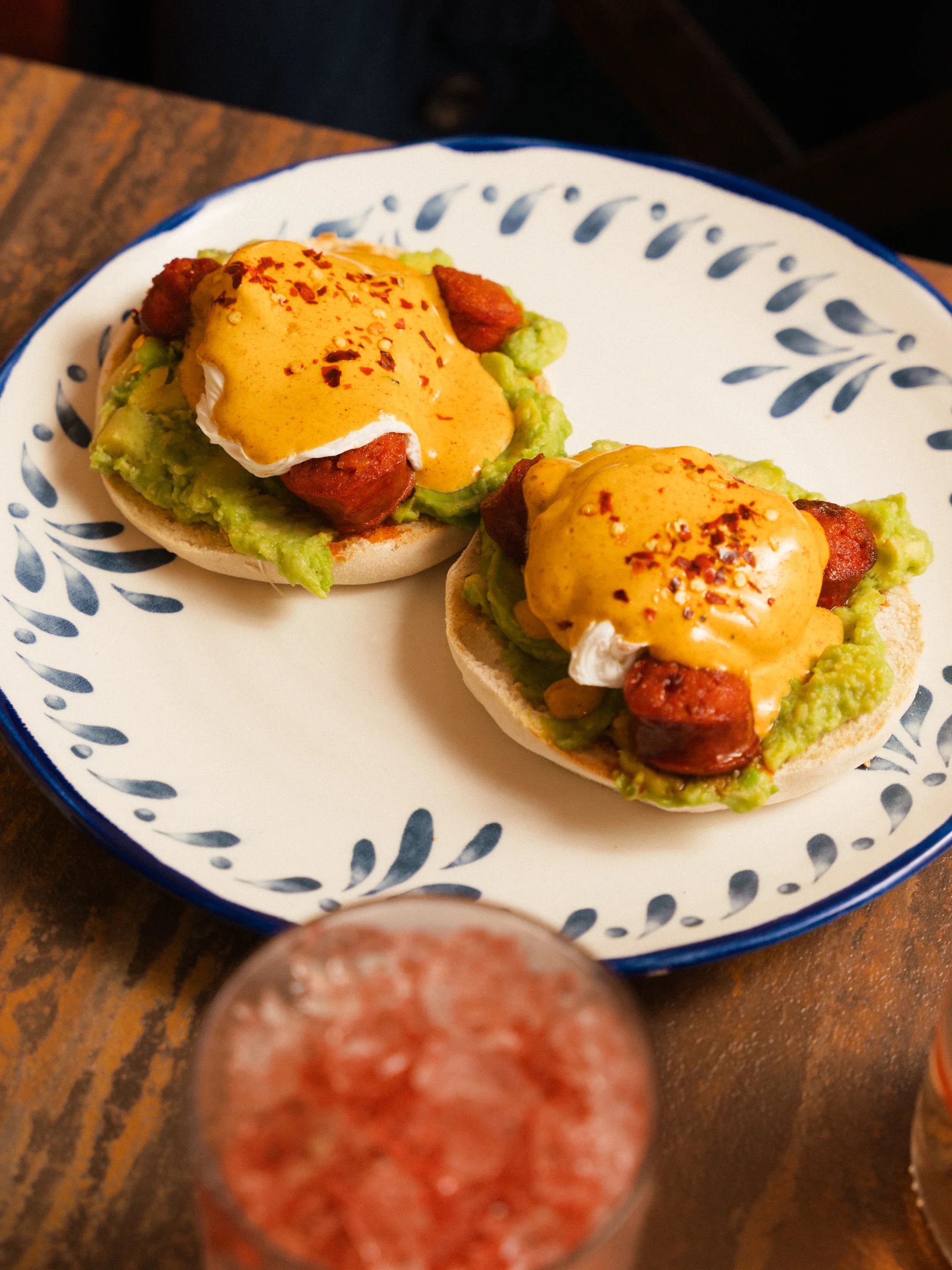Two open-faced chorizo and avocado sandwiches topped with poached eggs, spicy orange sauce, and red pepper flakes on a patterned plate