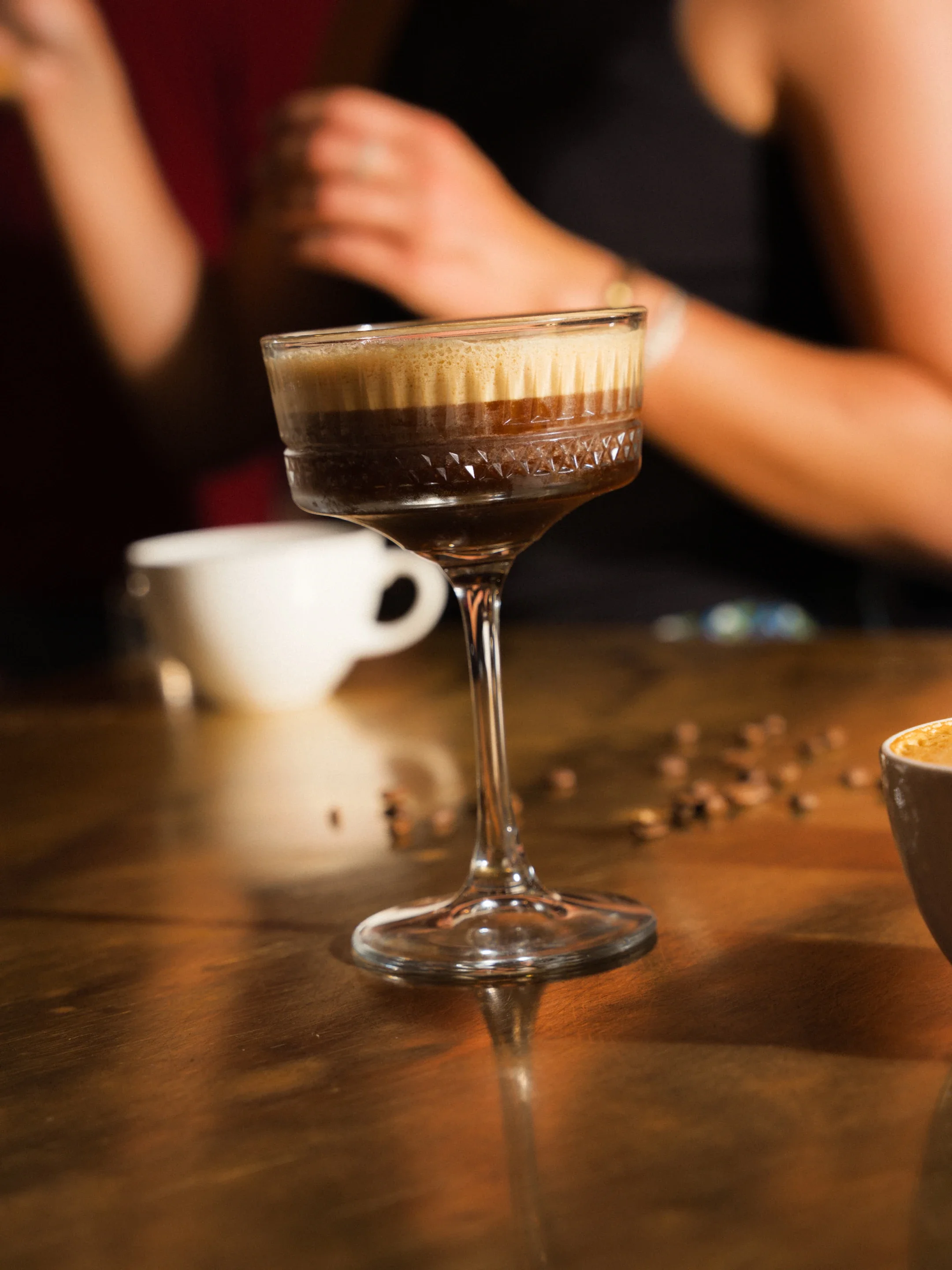 Layered espresso cocktail in a coupe glass on a wooden bar, with coffee beans and a small coffee cup nearby