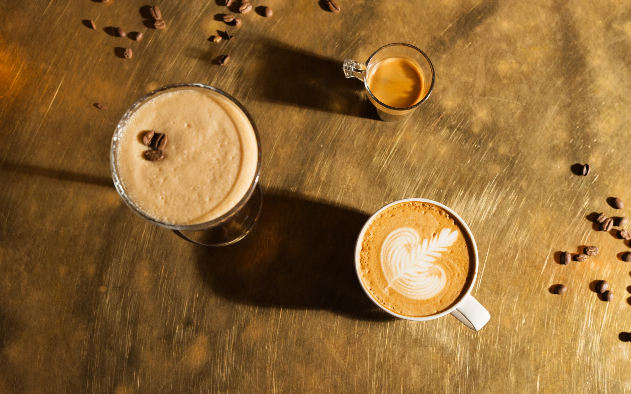 Overhead view of three coffee drinks on a wooden table, including a latte with leaf-shaped latte art, a frothy coffee, and an espresso, surrounded by roasted coffee beans.