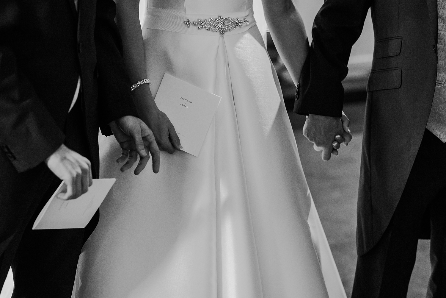  close up of groom and father of the bride holding the bride's hand. Can also see their names on the order of service. Black and white photo 