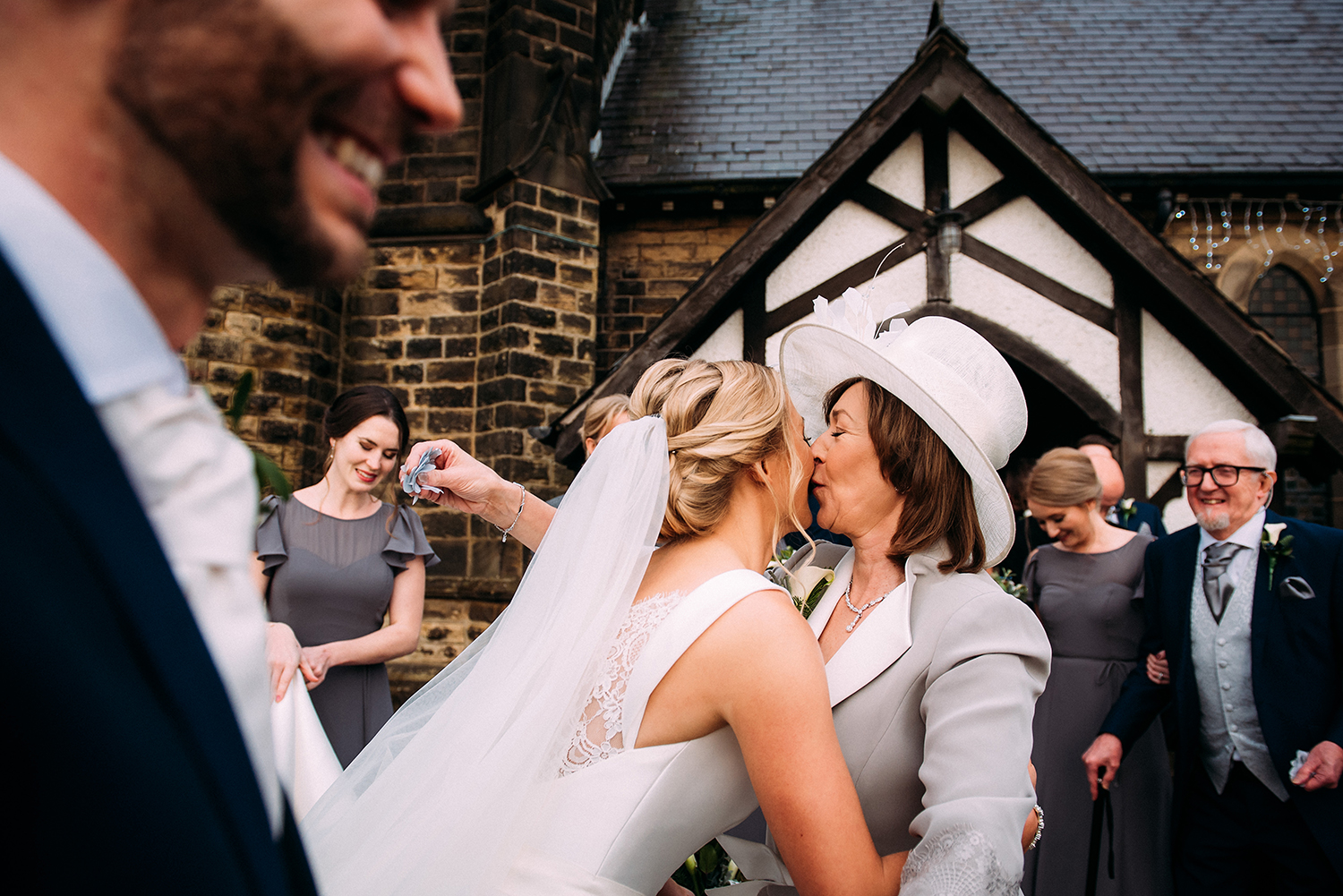  layered shot of bride and mother kissing, also has groom and bridesmaids in the foreground and background 