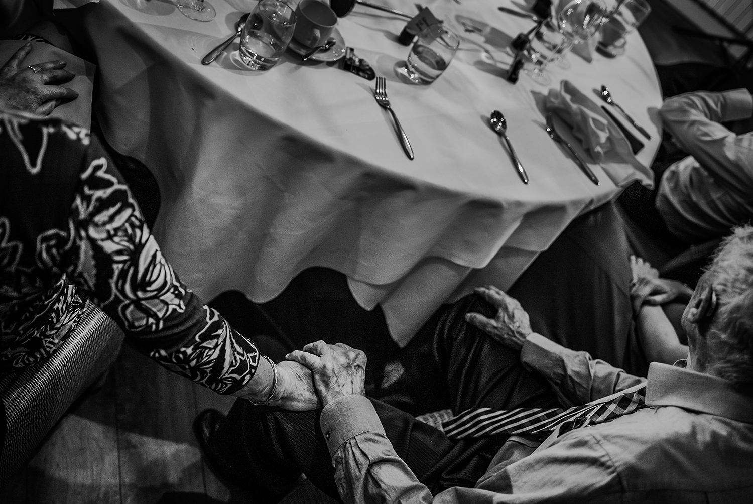  Black and white photo of the grandparents holding hands under the table during the speeches. 