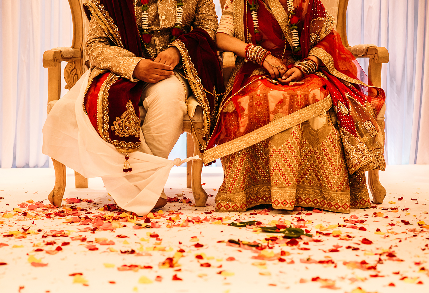  bride and groom from the waist down sat in ceremony chairs with traditional clothes. The floor covered in flower petals 