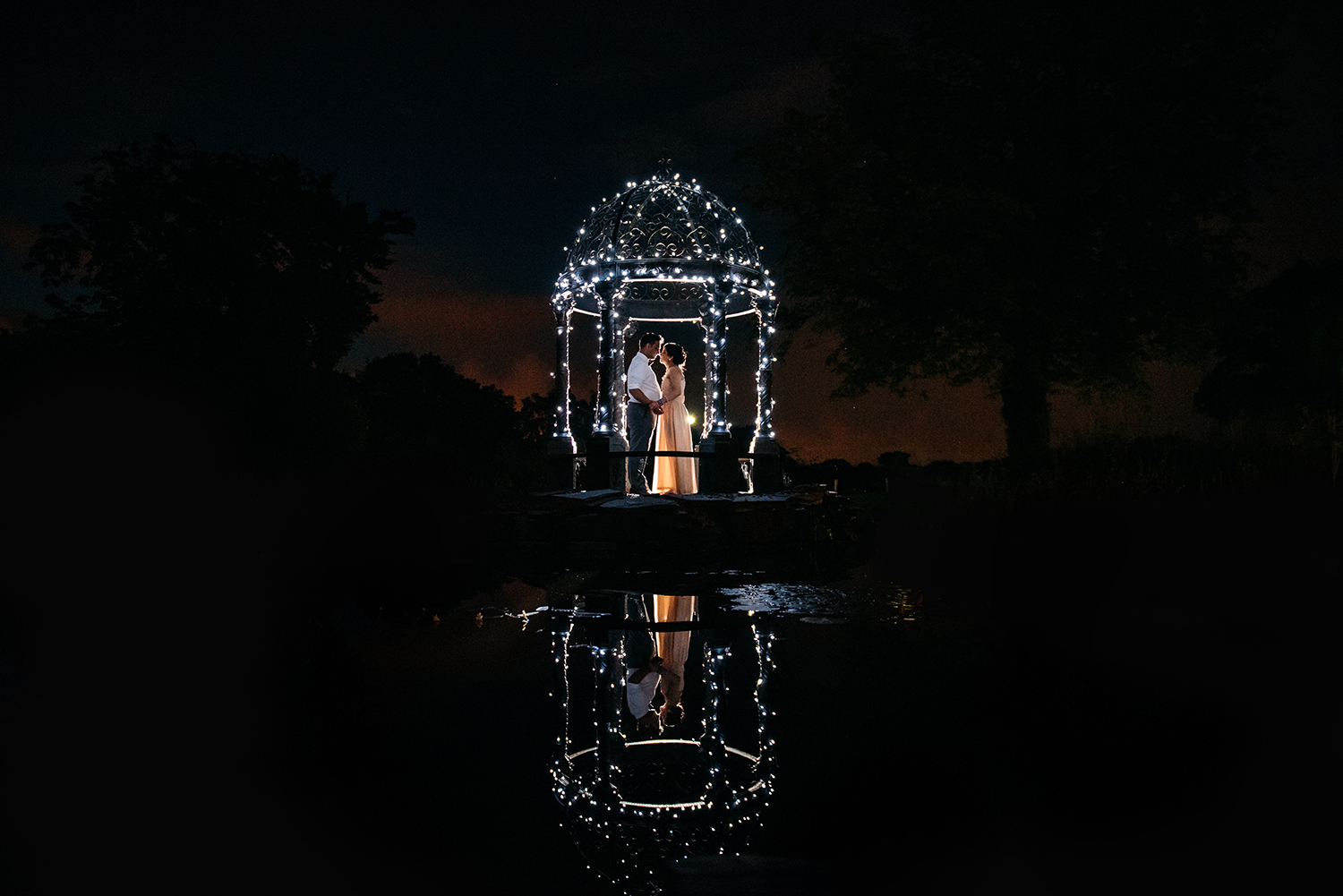  bride and groom together in a fairy lit gazebo at night with a reflection in the pond 