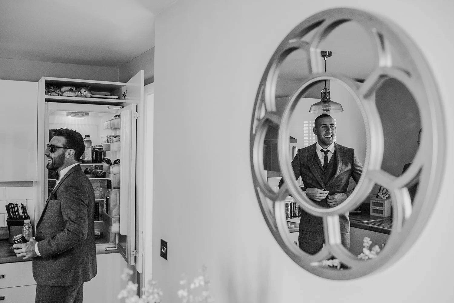 bw photo with the groom laughing as he gets a beer out of the fridge and a reflection of his best man laughing back at him.