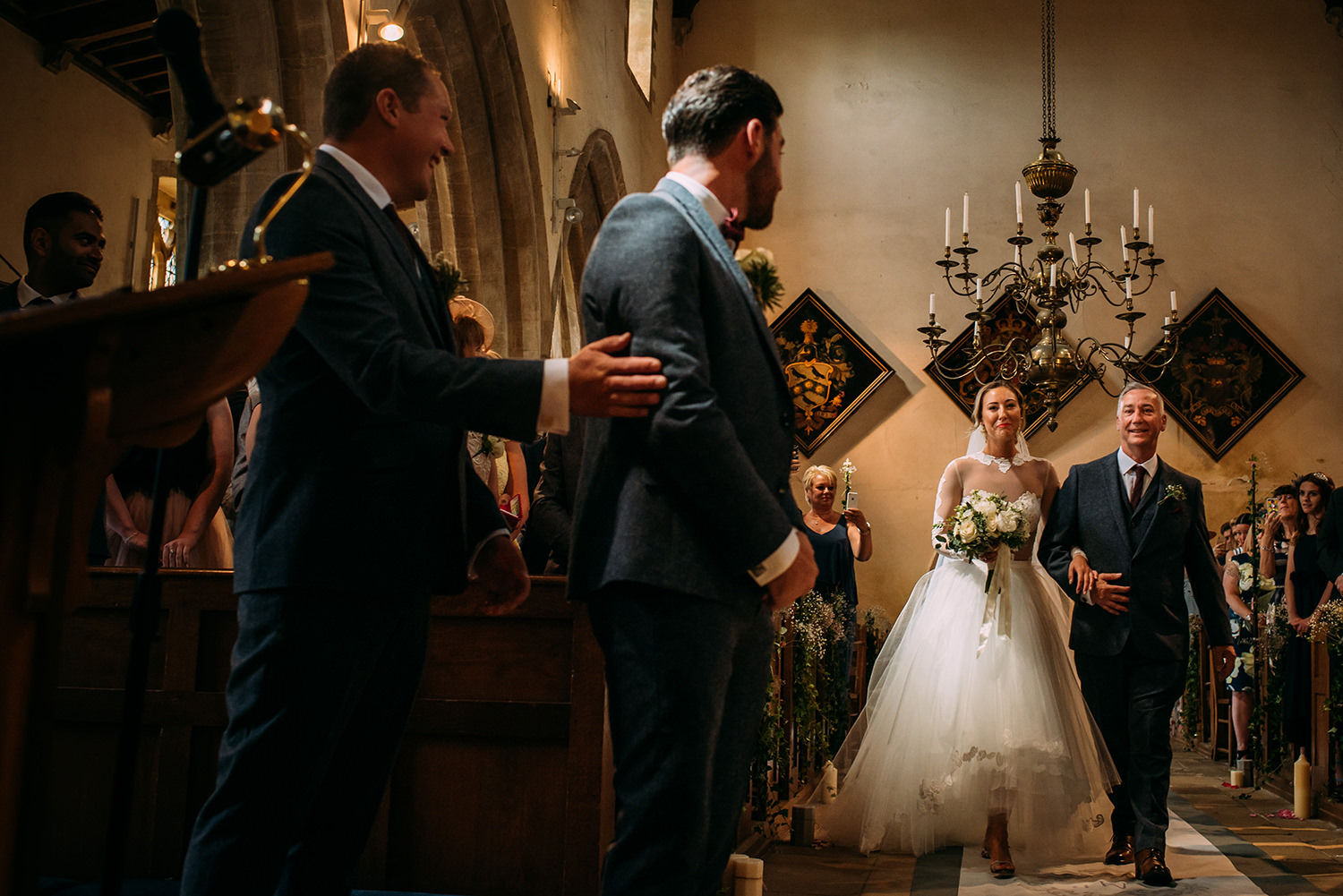  bride walking the aisle with her father, and bestman pats the grooms arm in the foreground 