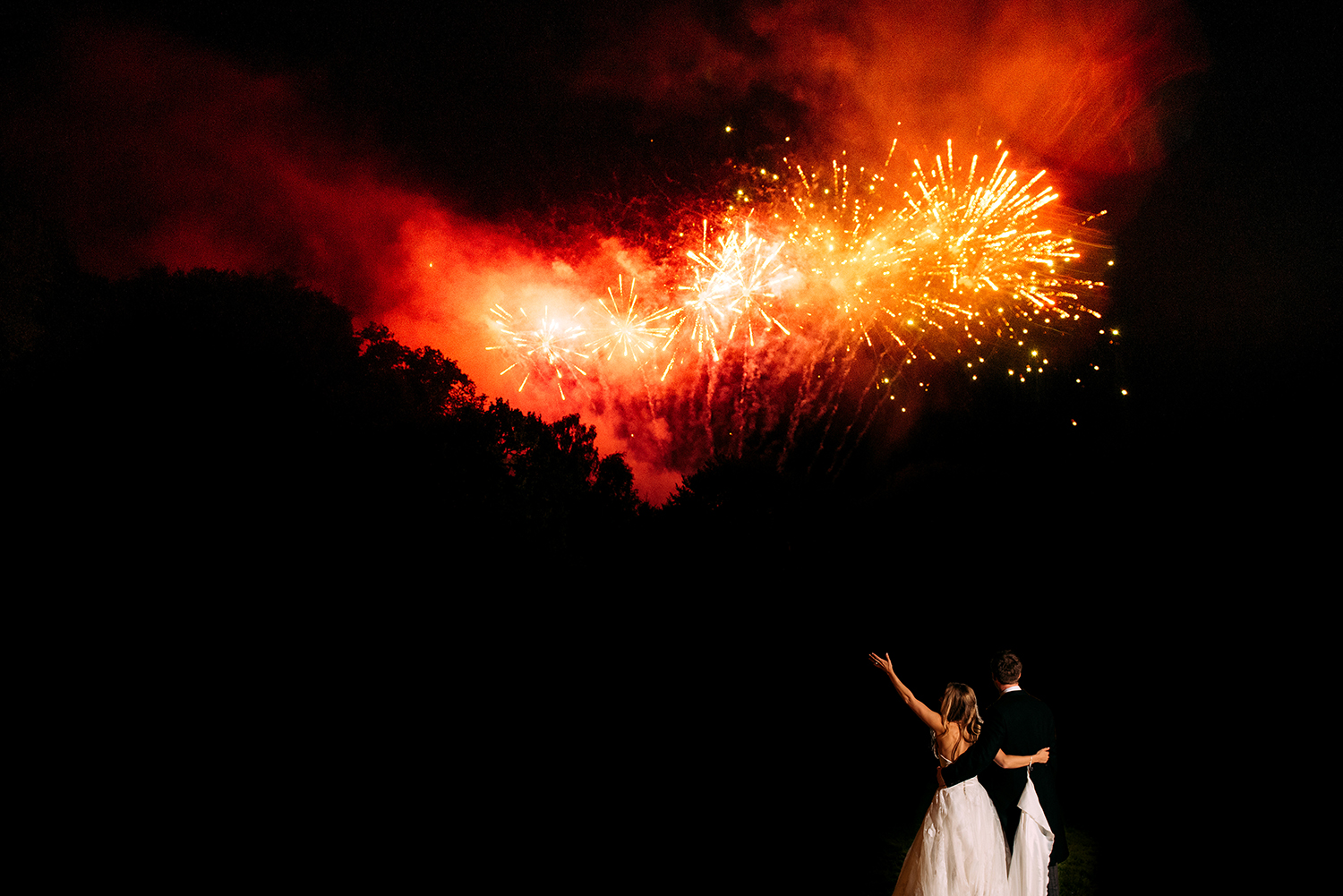  bride and groom. Bride lifts her arm in the air as fireworks go off 