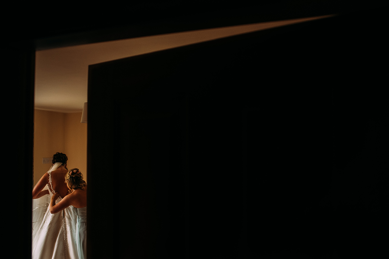 bridesmaid helping the bride into her dress, taken through a gap in the door