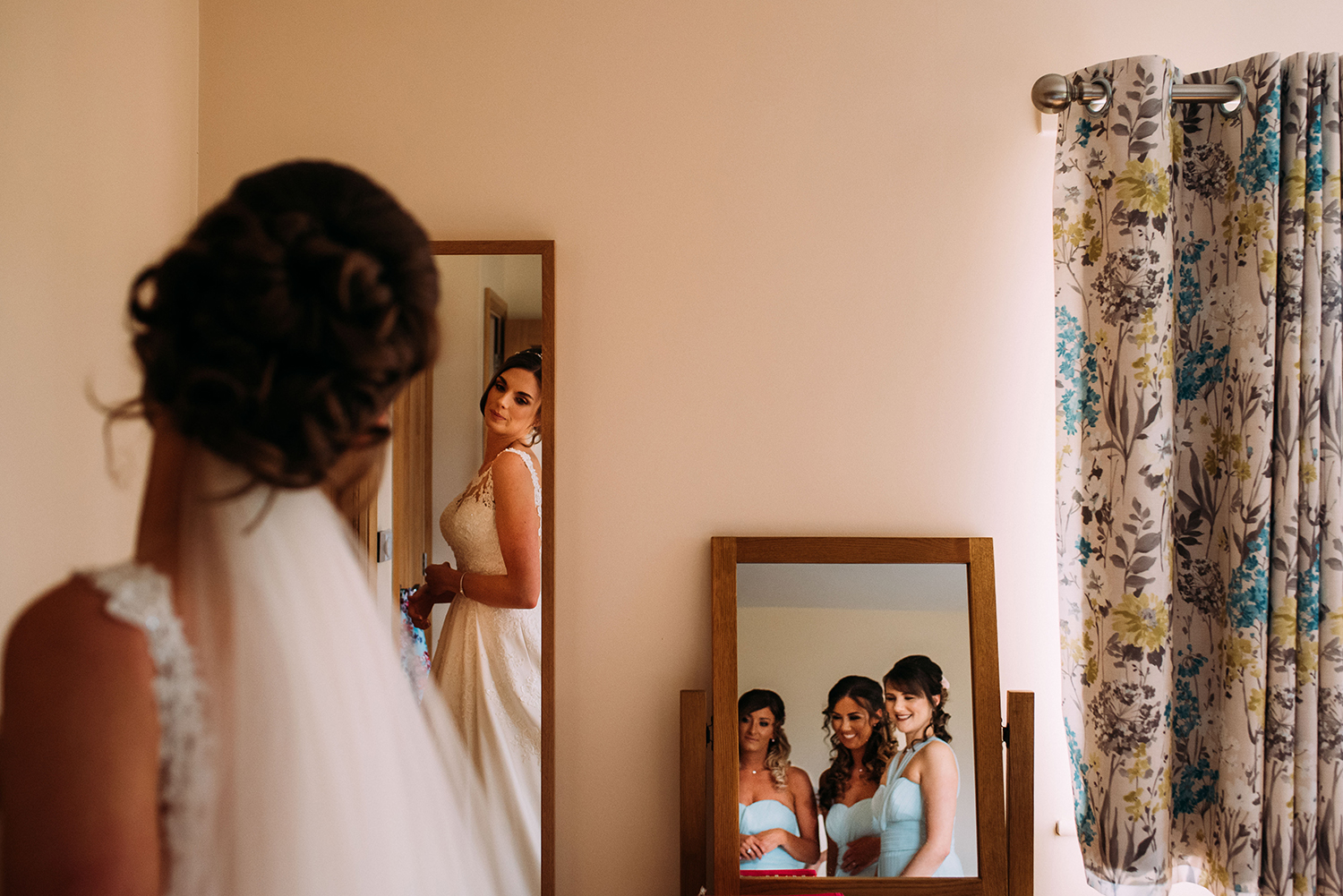 bride looks at her reflection in the mirror, the bridesmaids reflection can be seen in another mirror to the right