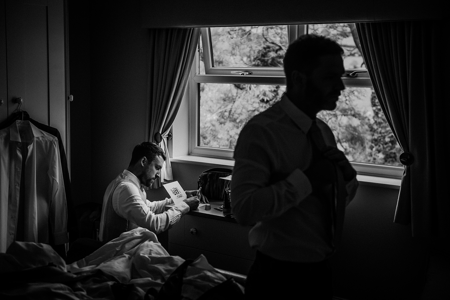 bw photo, groomsman writing a card with the silhouette of another doing his tie