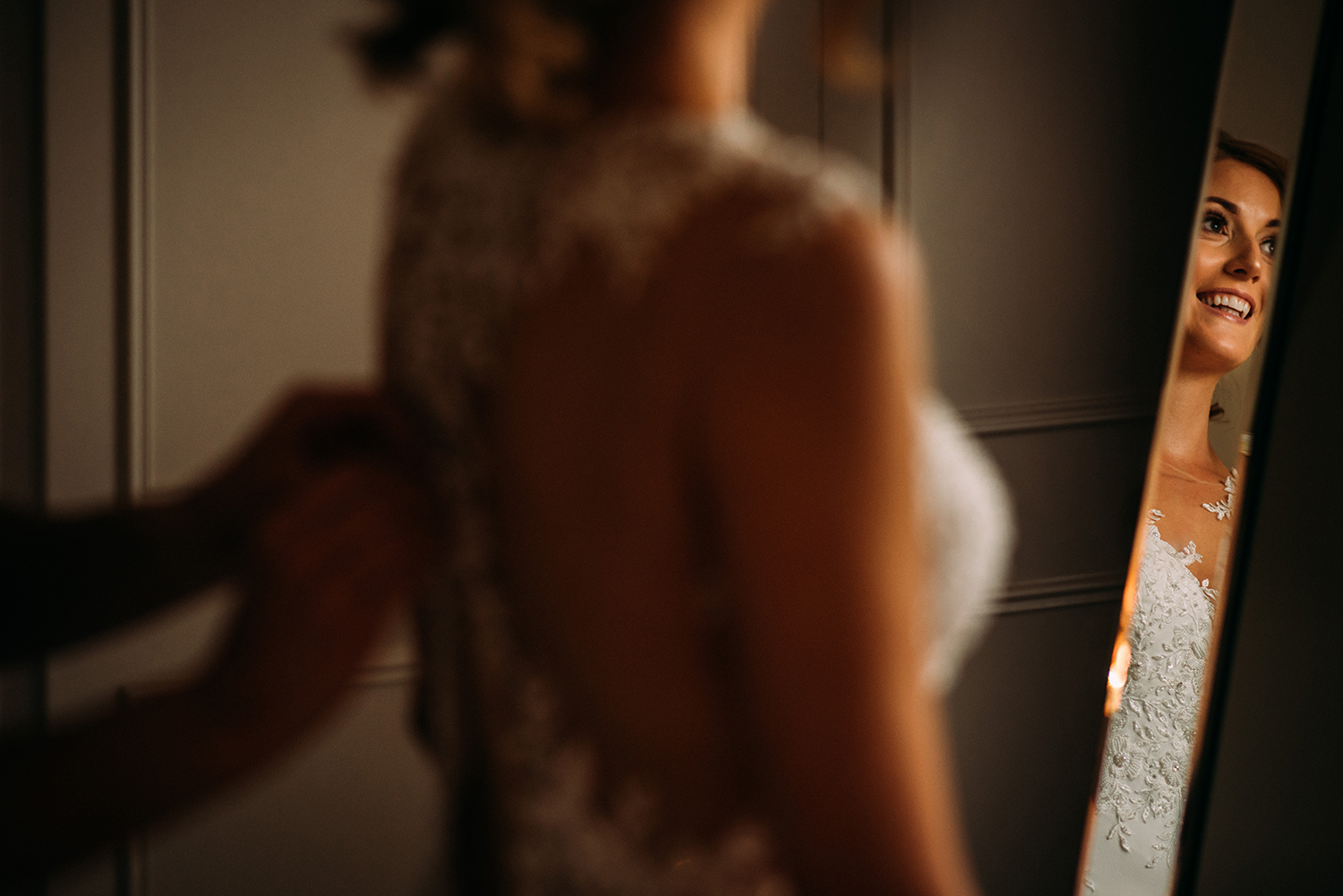 bride having dress fastened, slight reflection of her smile in the mirror