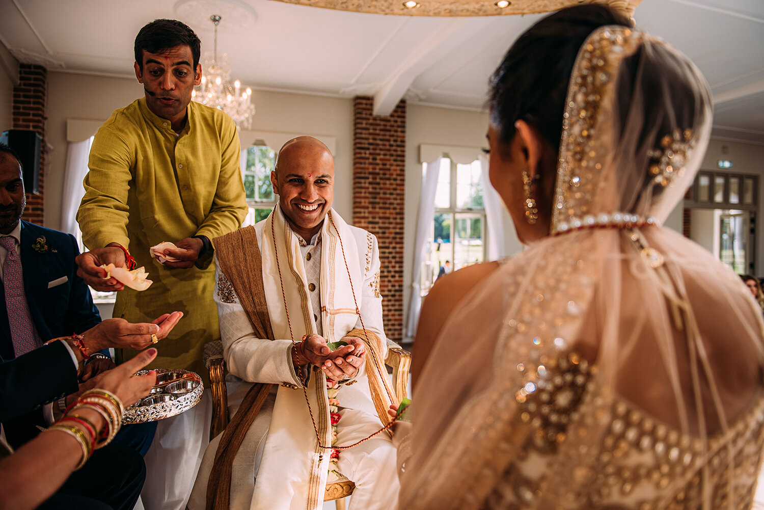  groom laughing and smiling at the bride on the mandap. Priest hands parent flowers to the left 