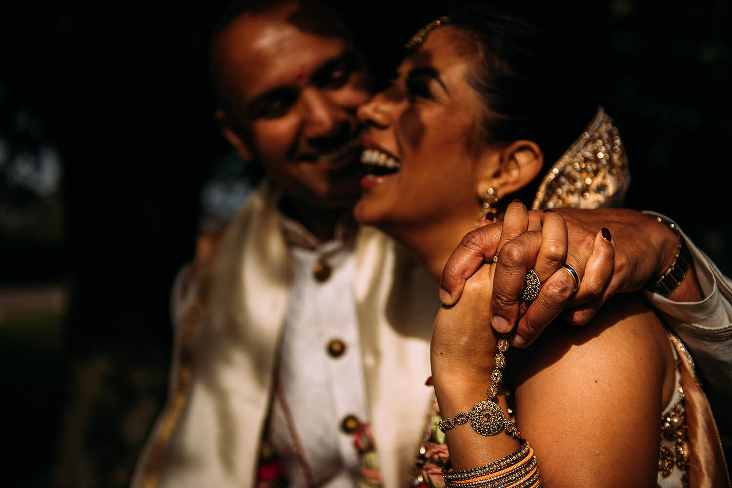  focus on bride and grooms hands holding on bride’s shoulder out of focus is the groom looking at the bride as she laughs 