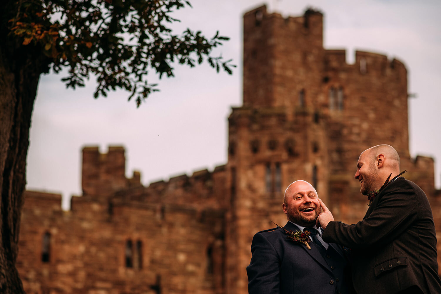  Two grooms laugh with each other in front of Peckforton Castle 