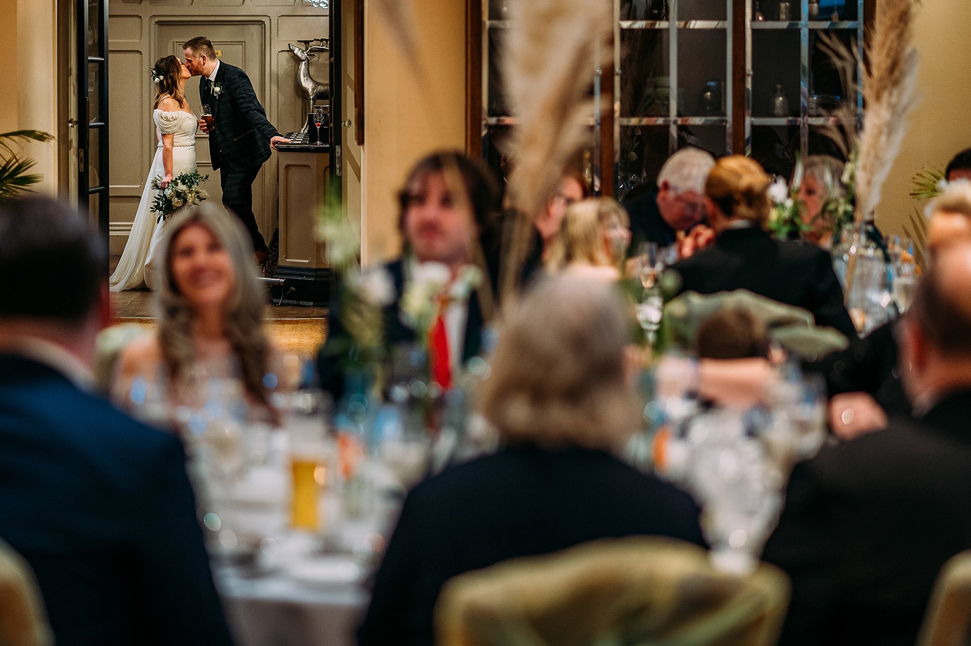  Taken through a room full of guests, bride and groom kiss in the background. 