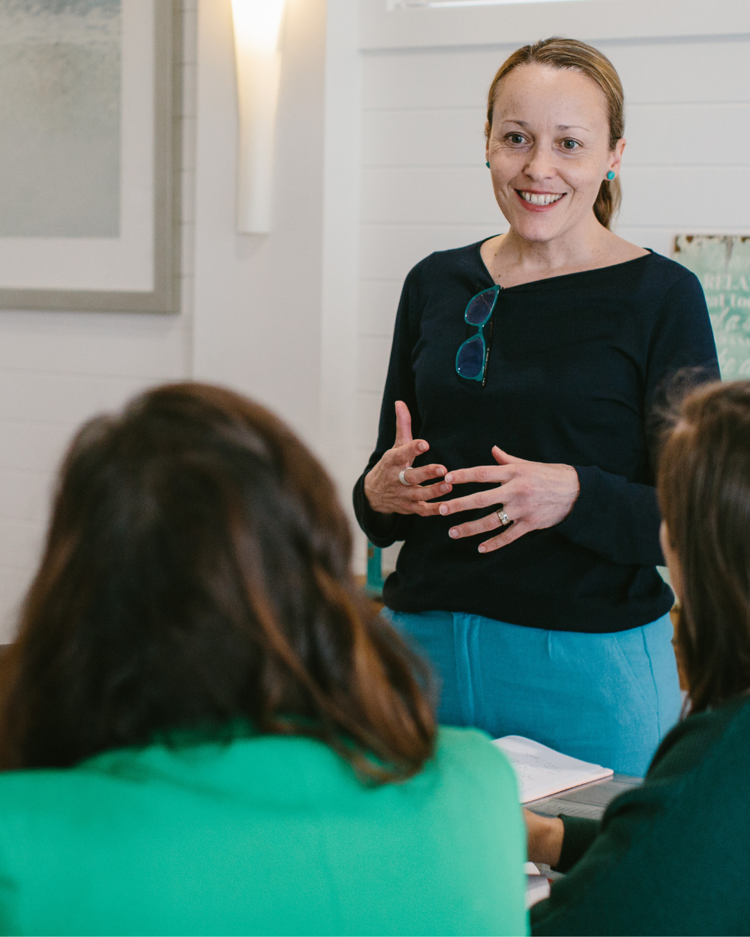 Erika McInerney stands in a room presenting a workshop to Australian small business owners and community organisations. She is wearing blue pants and a navy top and has her blue glasses tucked into her neckline.