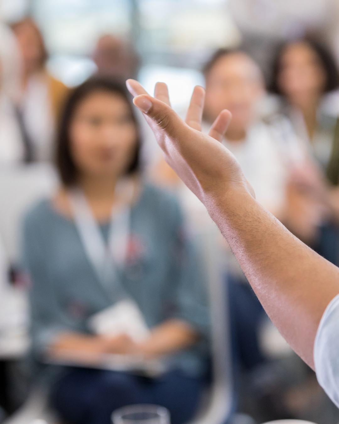 A hand in the foreground of a speaker, talking to a room of people wearing lanyards.