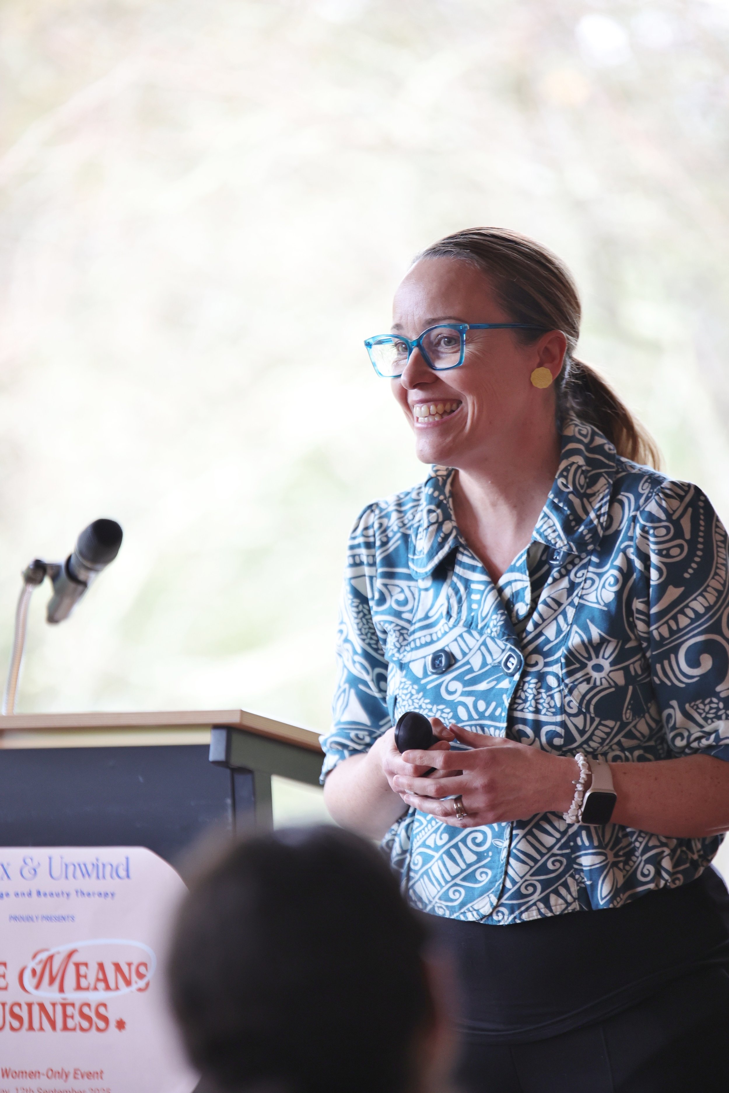 Erika McInerney presents a keynote on marketing at a female founders conference in the Yarra Valley. She is smiling at the audience and stands in front of a lectern. She is wearing a blue tribal print shirt and has blue glasses.