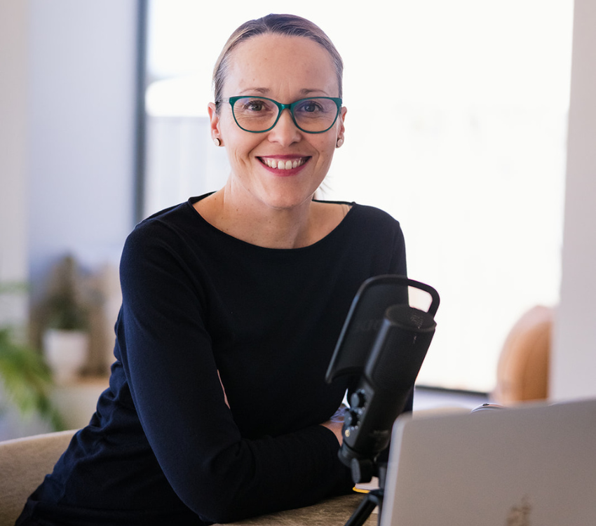 Erika McInerney sits in front of a microphone and laptop, she's smiling and has her hair pulled back. She's wearing a long sleeve navy tee and green glasses.