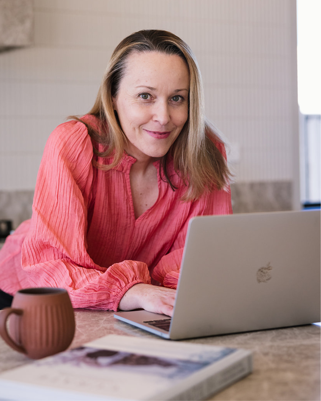 Erika McInerney sits at a laptop, smiling at the camera, wearing a pink long sleeve shirt. Erika provides marketing, content and customer experience strategy to small business and community organisations across Australia.