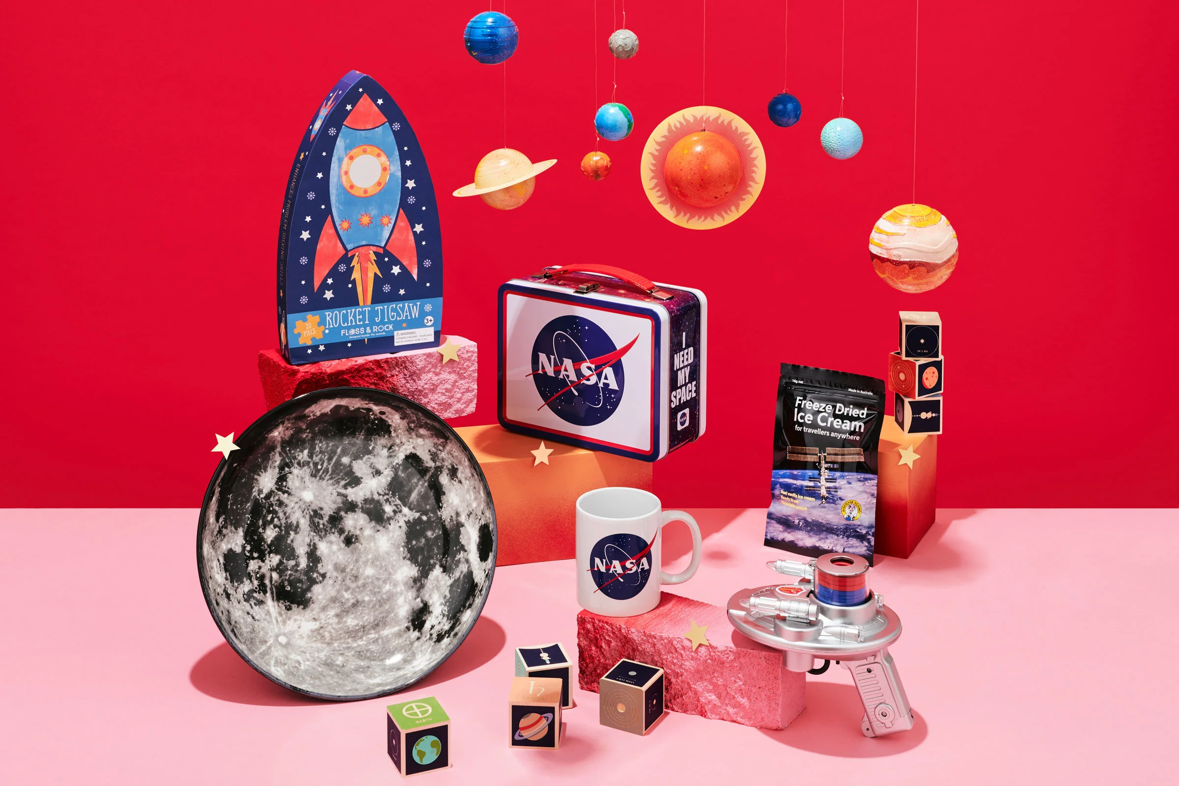 Colorful space-themed decorations including planets, moon, rocket jigsaw puzzle, NASA logo, ice cream, toy gun, and various blocks with space and Earth designs, set against a vibrant red background.