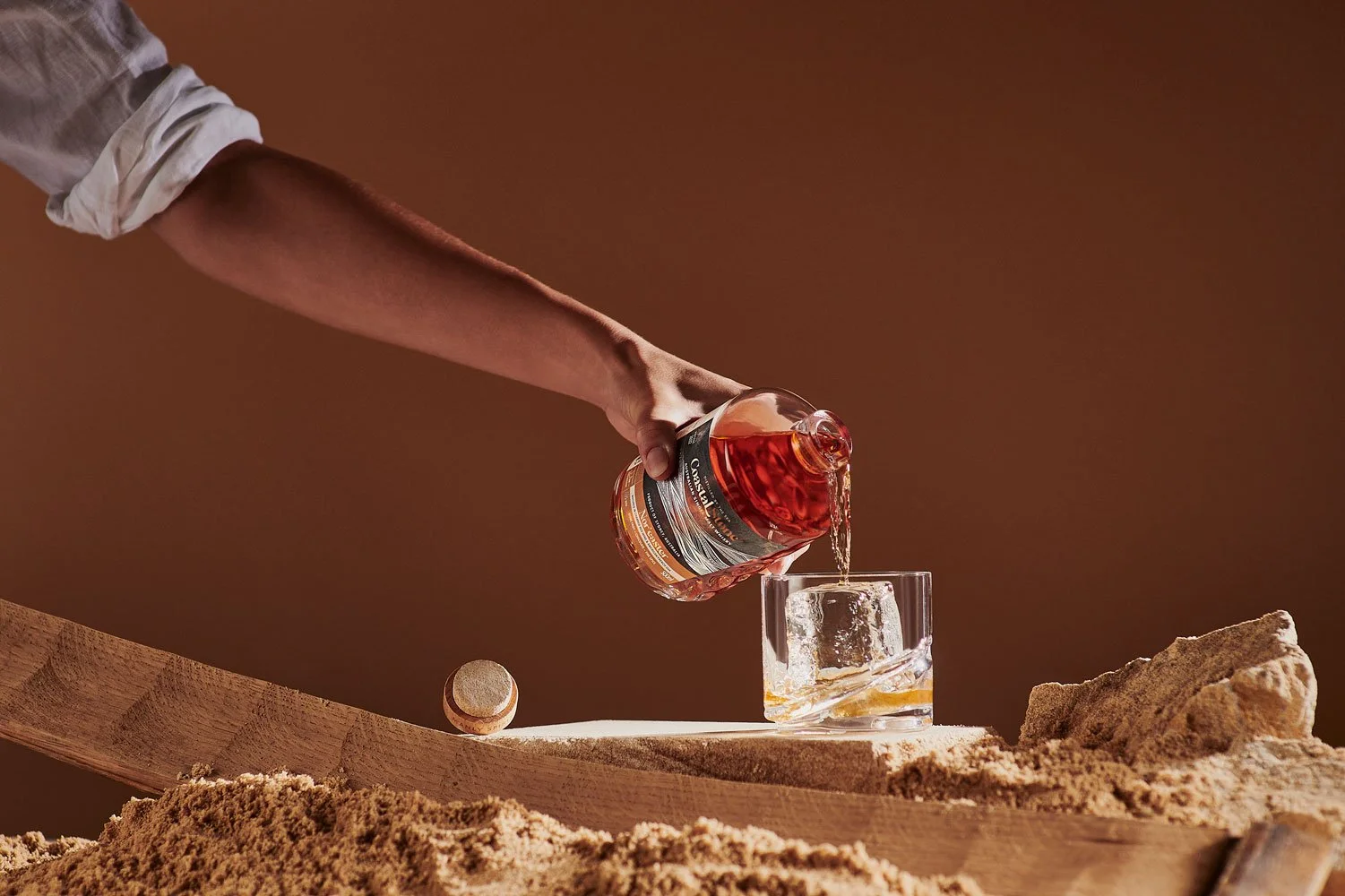 A person with a rolled-up sleeve is pouring a bottle of whiskey into a glass on a wooden surface, with a sandy background and a small wooden ball nearby.
