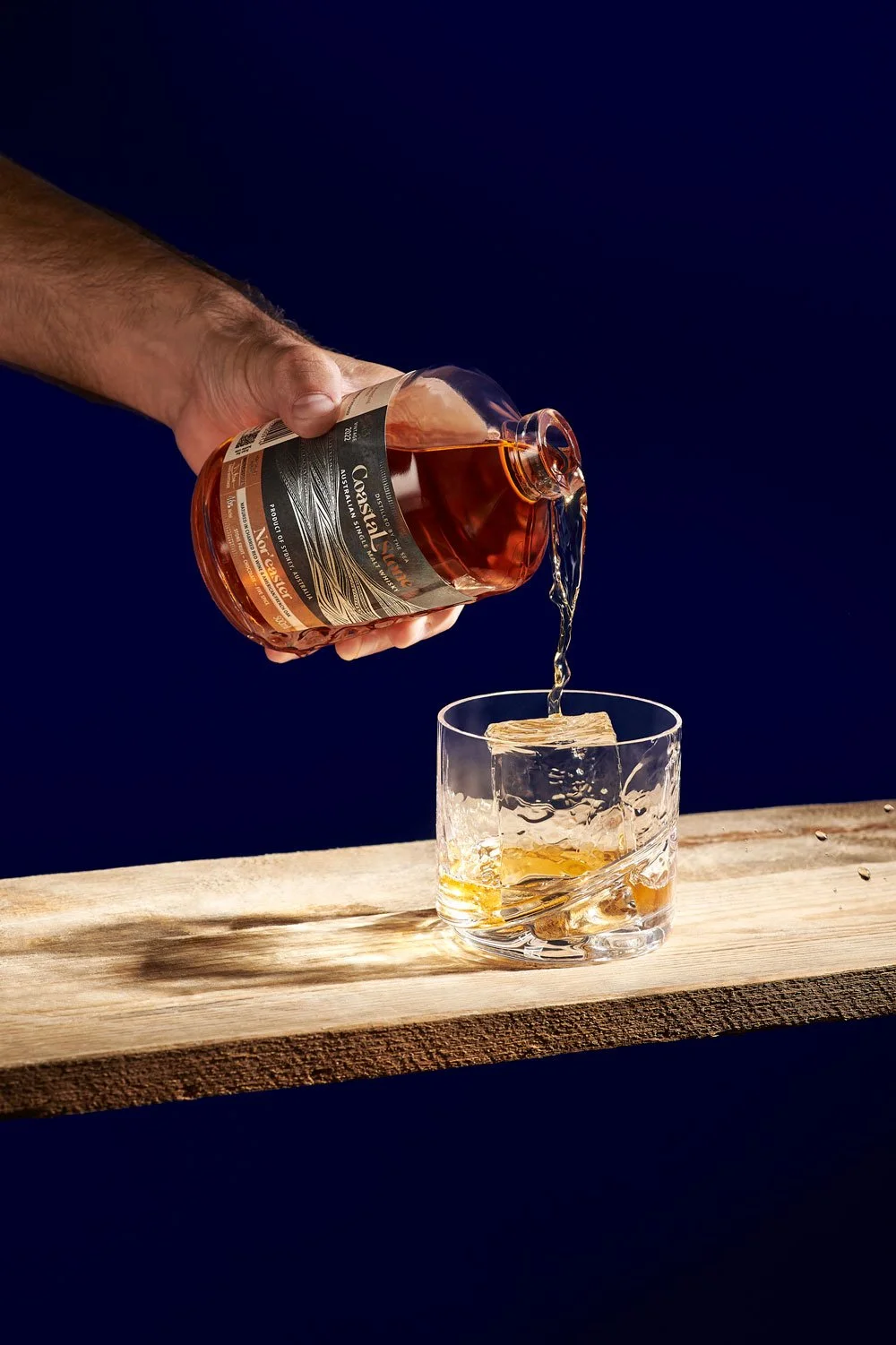A person pouring whiskey from a glass bottle into a glass with ice on a wooden surface against a dark blue background.