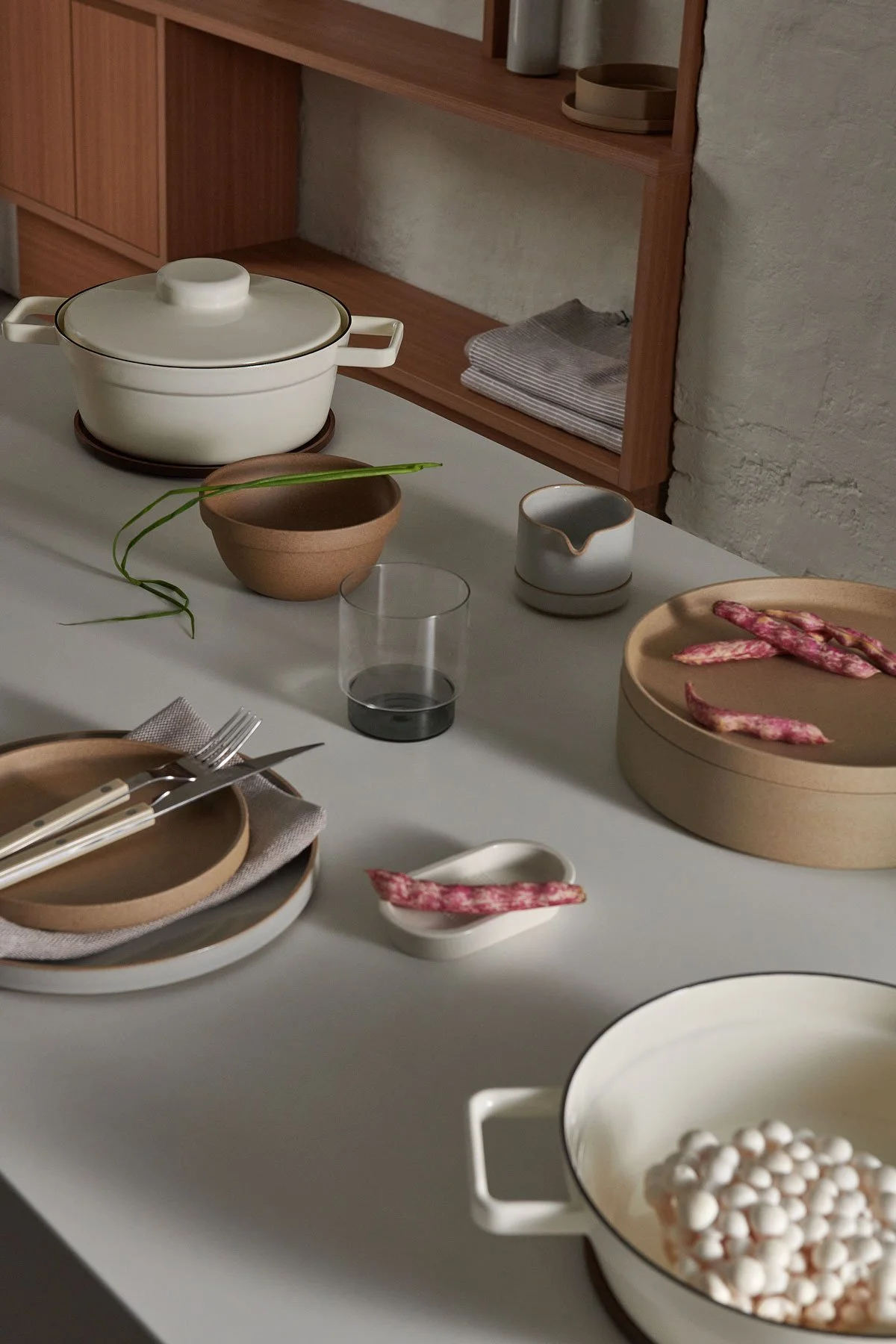 A minimalist dining table set with dishes, utensils, a potted plant, and a bowl with pink and white chips, featuring a wooden cabinet in the background.