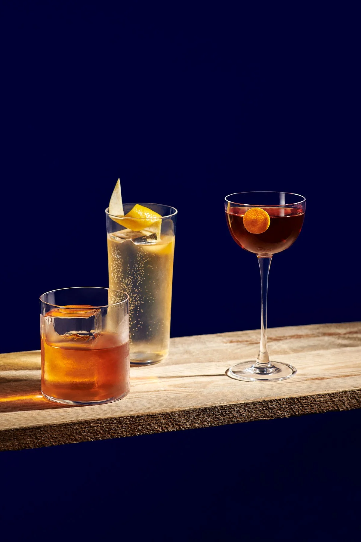 Three cocktails on a wooden board against a dark background. The first is an orange-colored drink with a slice of orange, the second is a clear beverage with lemon and lime slices, and the third is a dark red drink with a small round garnish.