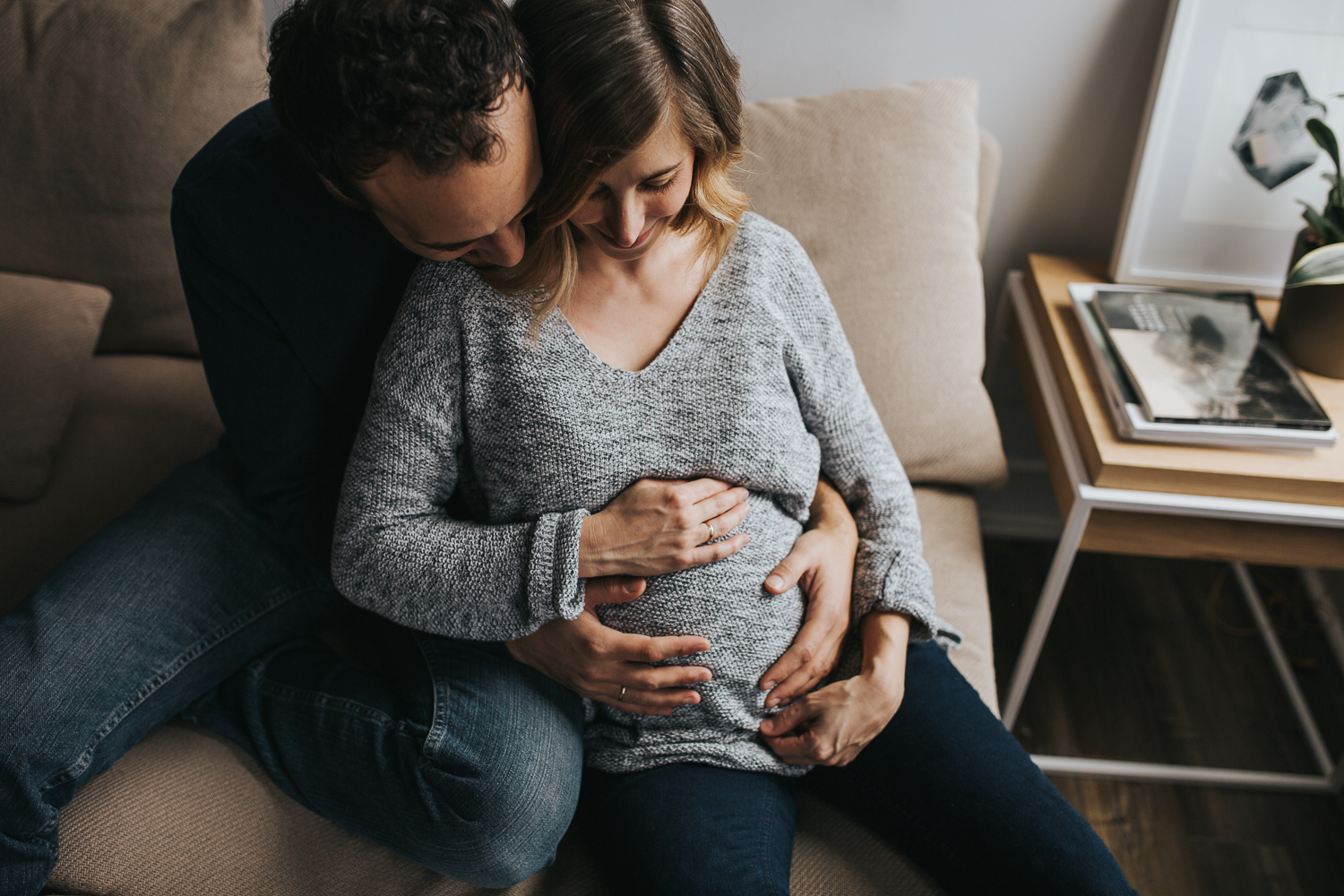 husband embraces pregnant wife on couch, hands on baby bump - newmarket in-home family photos 