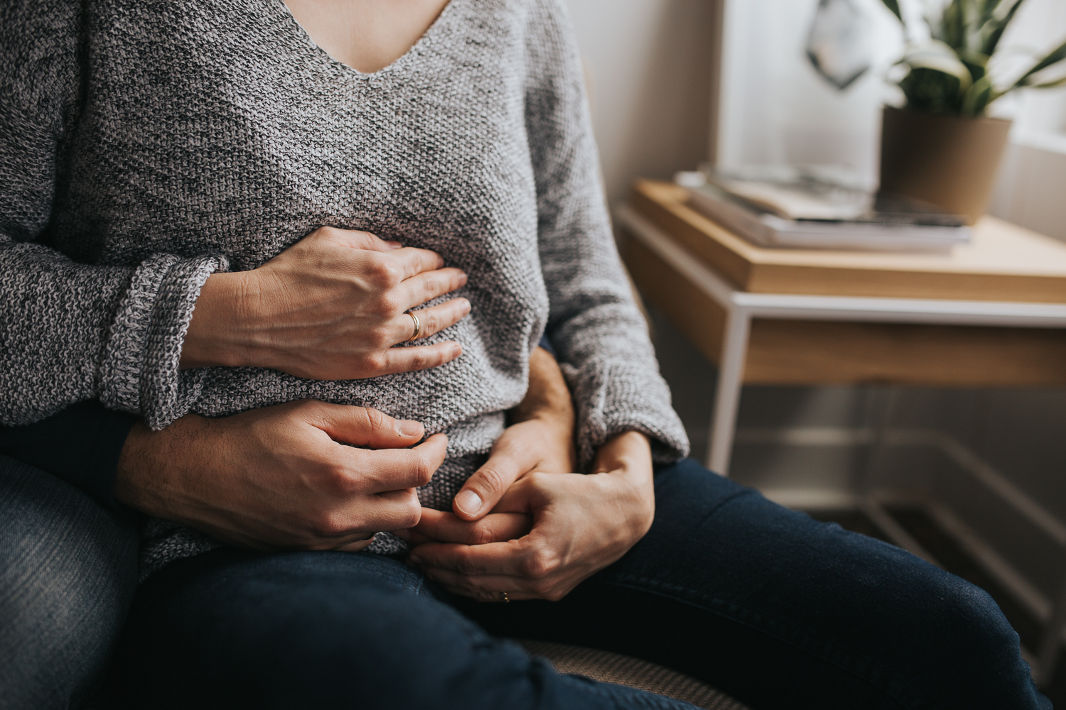 husband embraces pregnant wife on couch, hands on baby bump - newmarket in-home family photography