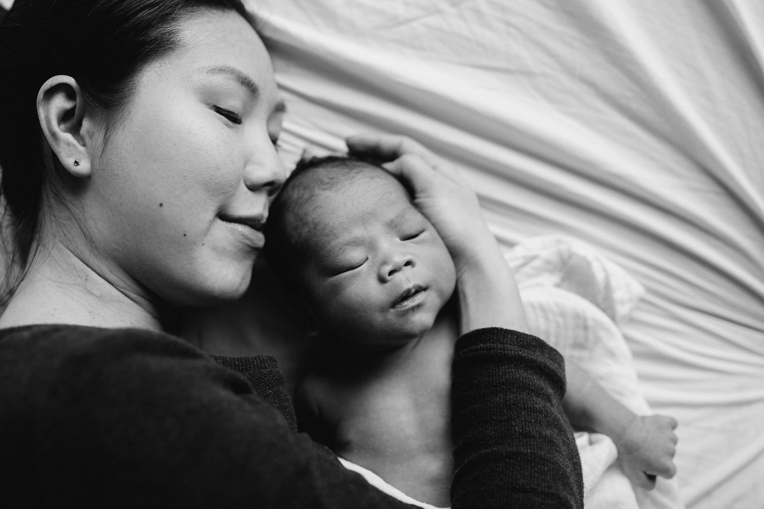 mom snuggling 2 week old baby boy with dark hair while lying on bed - Stouffville in-home photography