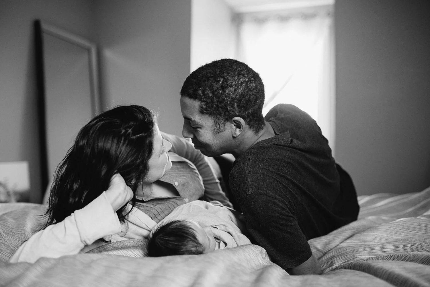 husband and wife lie on bed with 2 week old baby girl between them, smile at each other - Newmarket in-home photography session