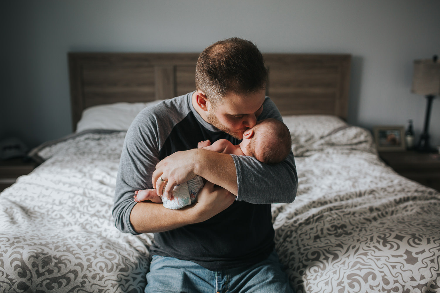 new dad sitting on bed holding and kissing 6 week old baby daughter - Barrie lifestyle photos