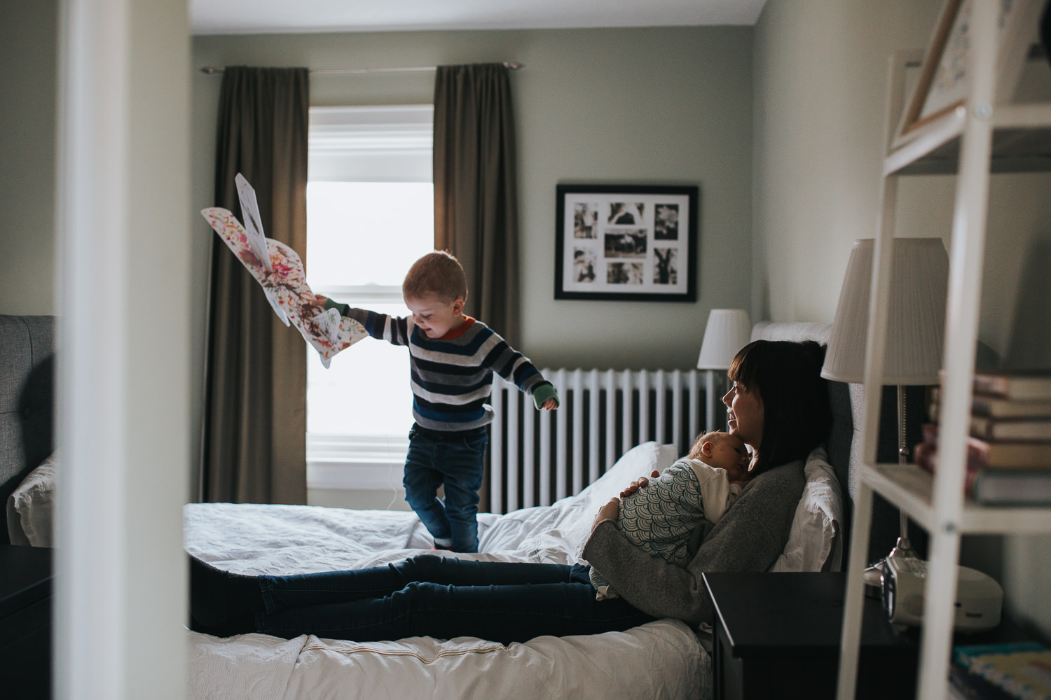 mom sitting on master bed holding baby girl, while toddler son stands on bed flying toy airplane - Markham lifestyle photography