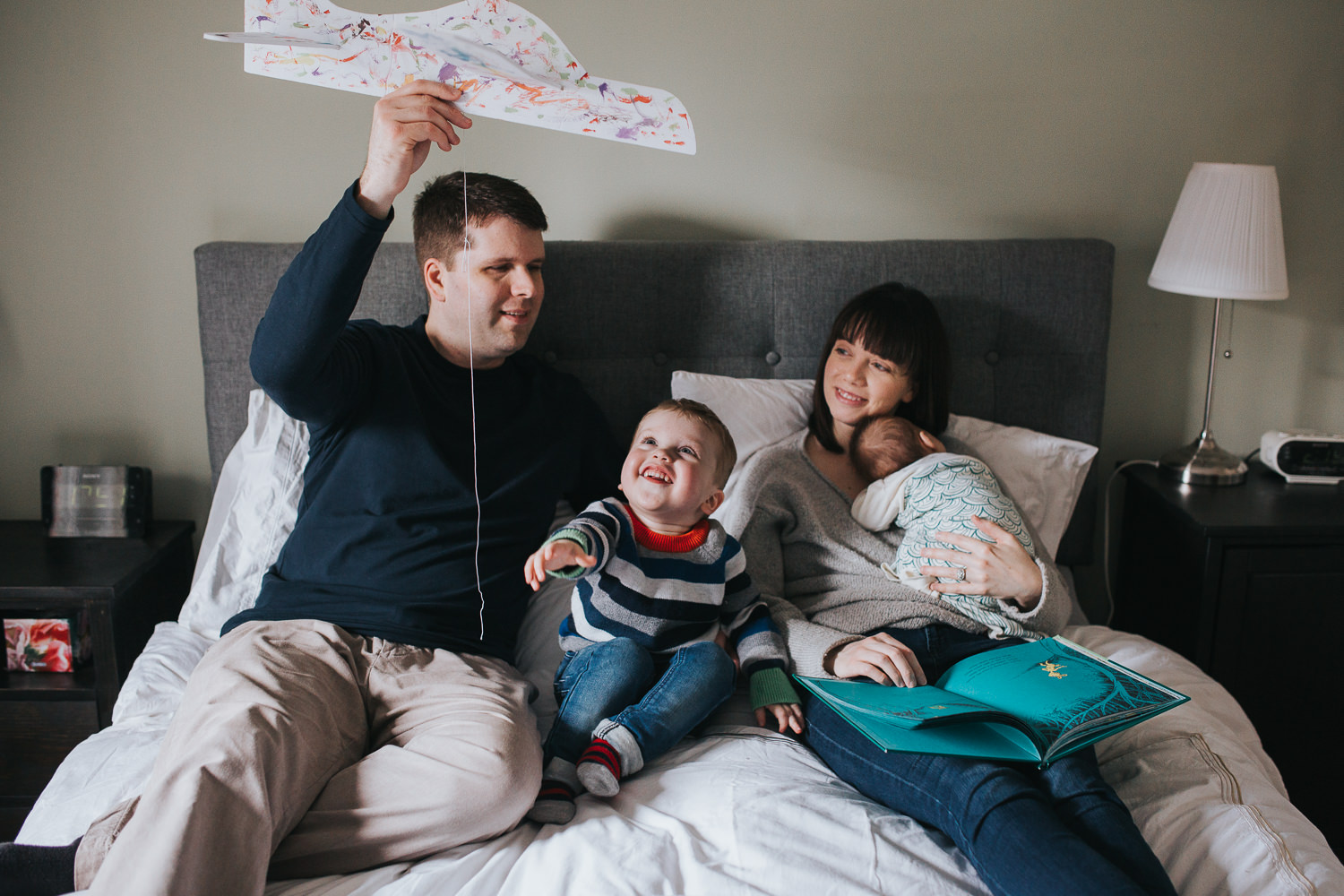 new family of 5 sits on bed, dad playing with toddler boy and mom holding baby girl - Newmarket in-home photography