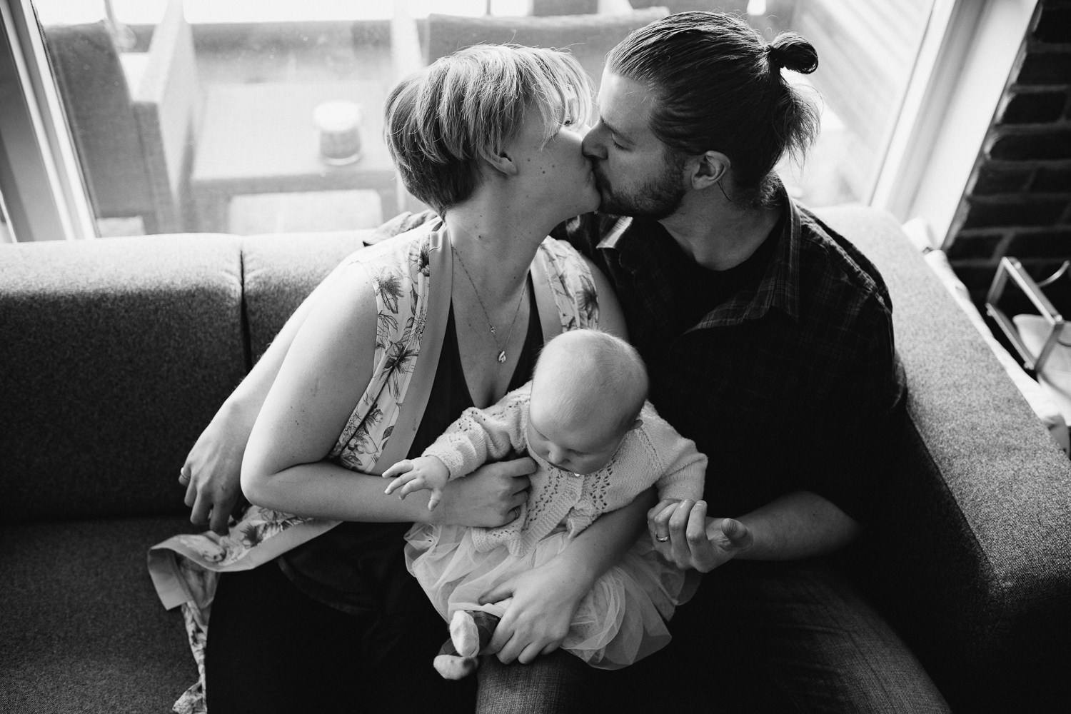 family of 3 sitting on couch, husband and wife kissing with 5 month old baby girl between them - Newmarket lifestyle photos