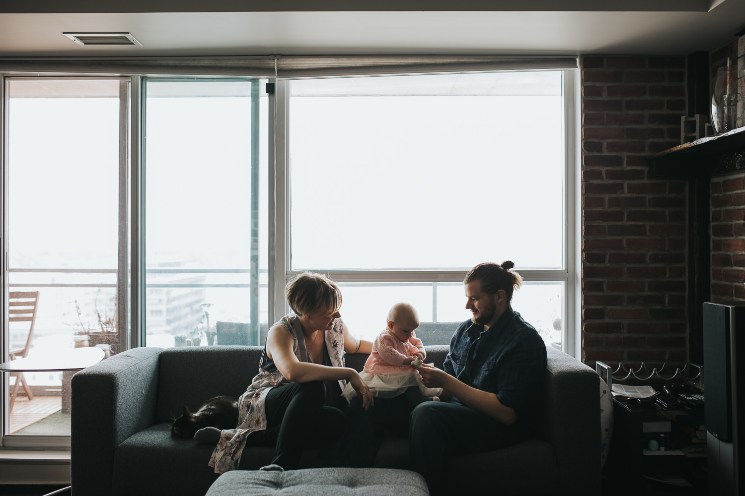 family of 3 sitting on couch in front of window as parents play with 5 month old baby girl - Barrie in-home photos