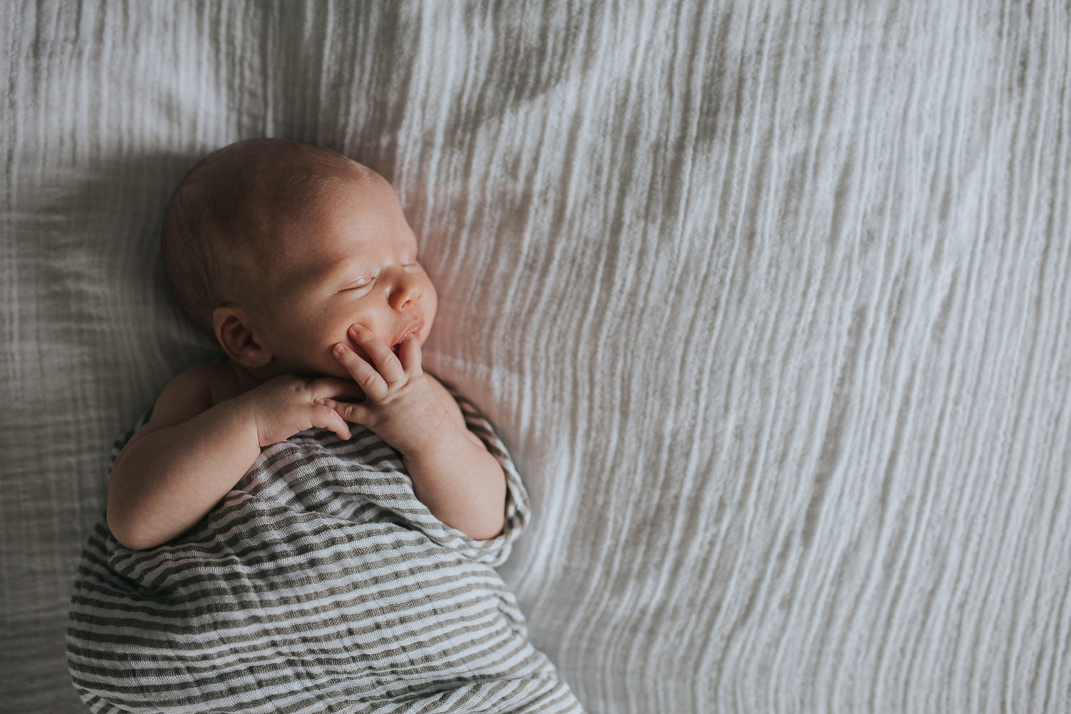 2 week old baby boy asleep, lying on bed in striped neutral swaddle with hand in mouth - Markham lifestyle photography