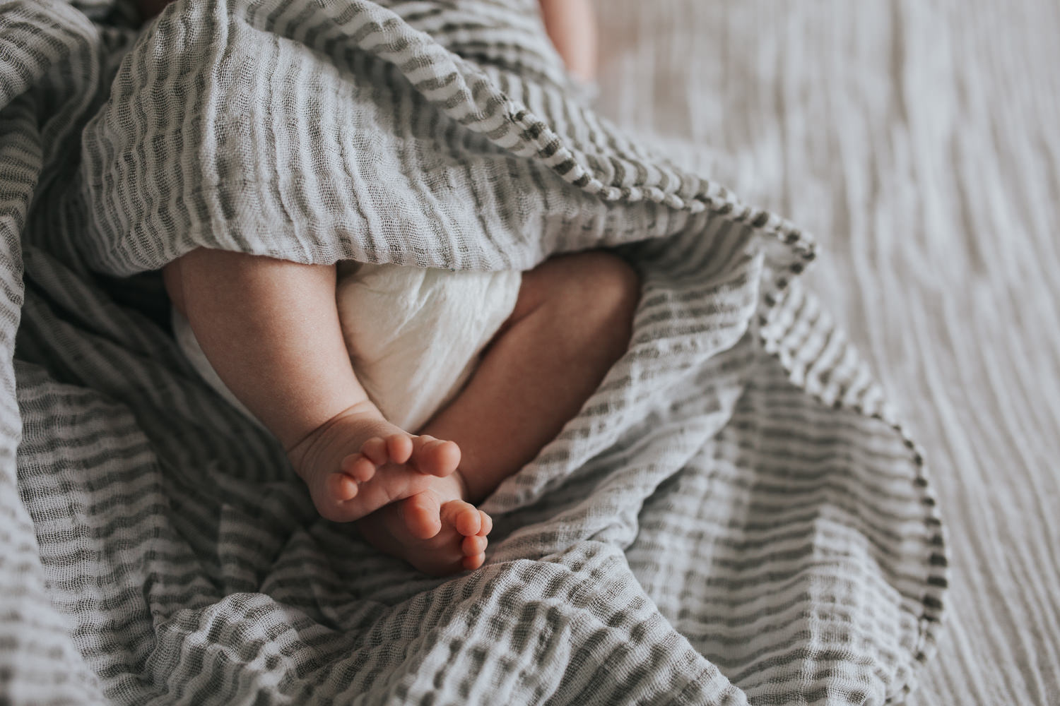 close up of 2 week old baby boy's feet, lying on white bed in striped neutral swaddle - Barrie lifestyle photography