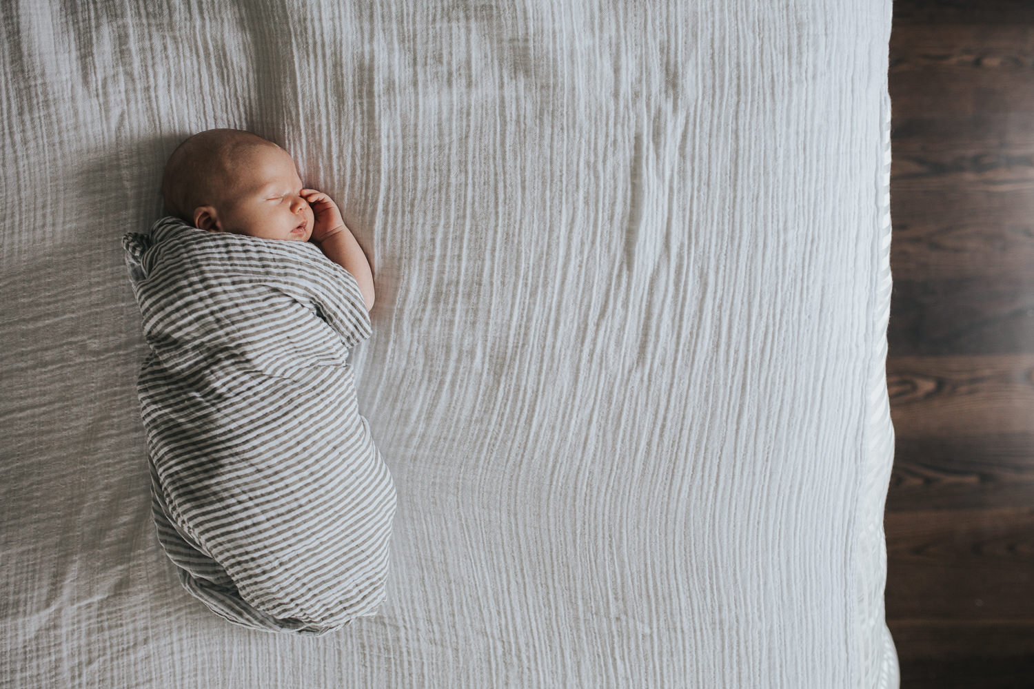 2 week old baby boy asleep, lying on bed in striped neutral swaddle with hand resting against face - Newmarket in-home photography
