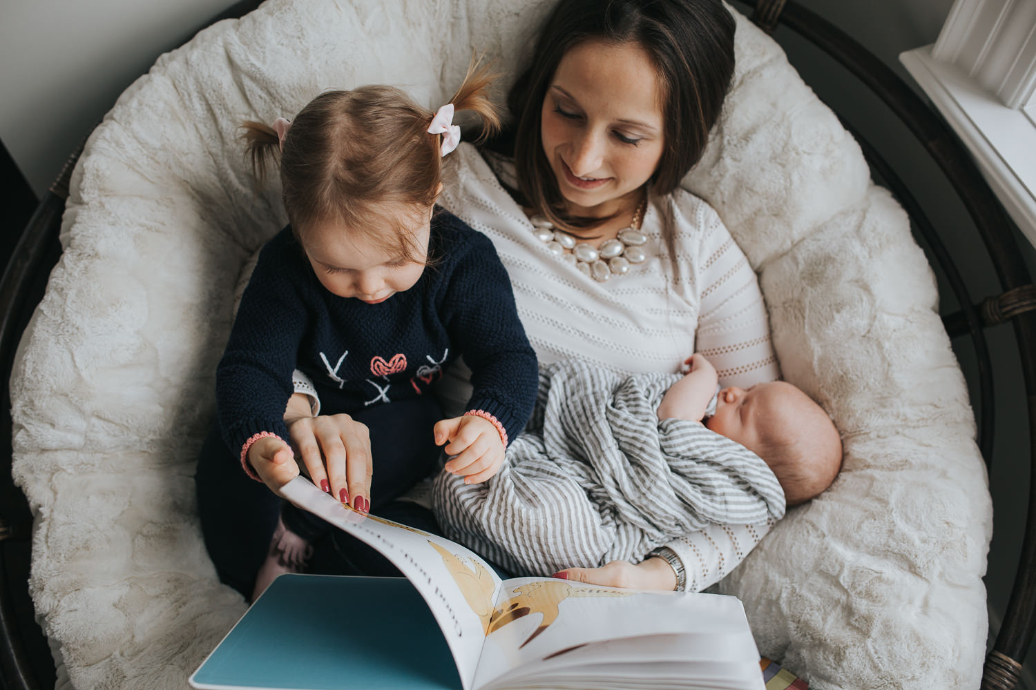 mom sitting in chair holding 2 week old baby boy and with 2 year old toddler girl in lap, reading book - Stouffville in-home photography