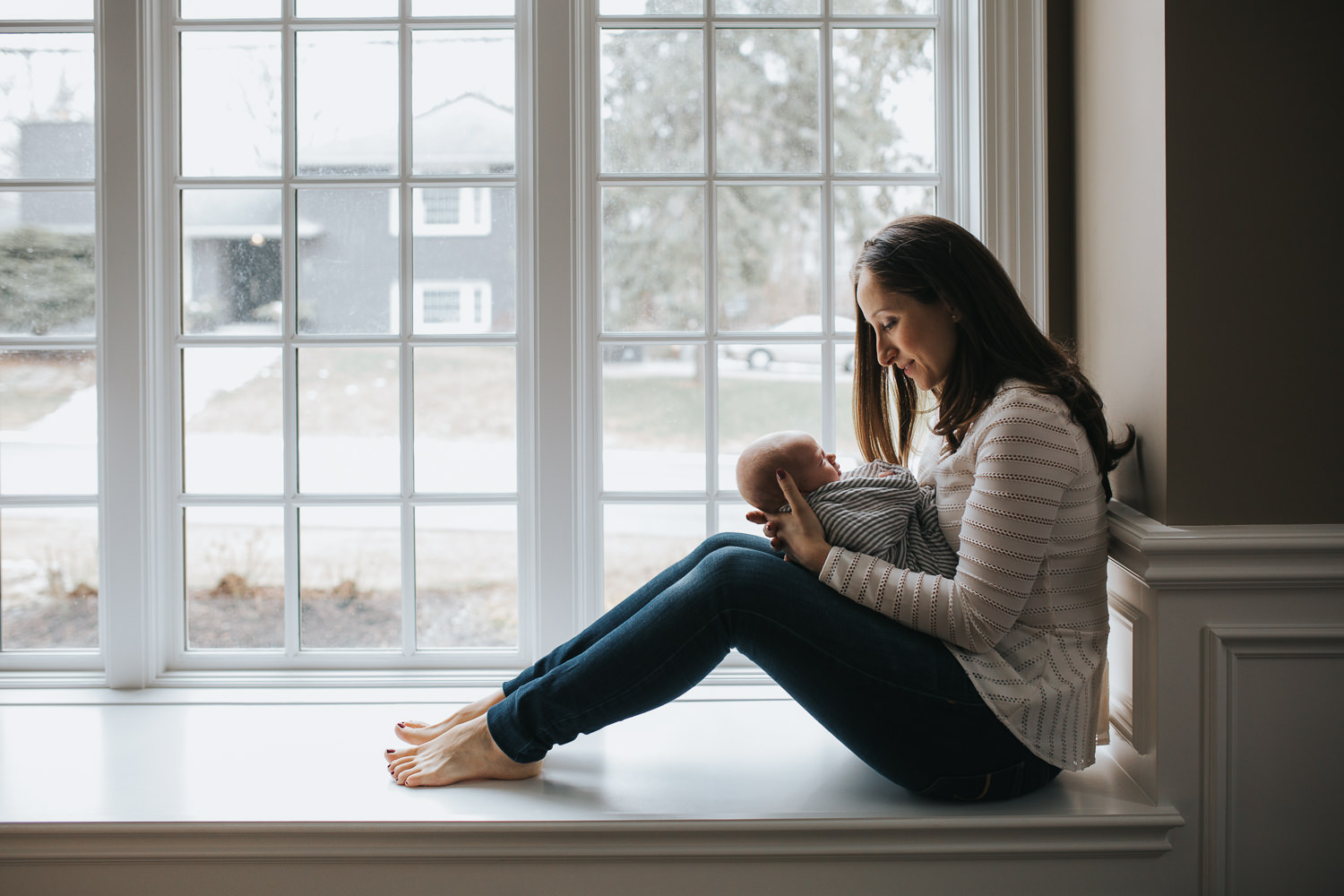 mom sitting on window seat holding and looking at sleeping, swaddled 2 week old baby boy - Stouffville lifestyle photos