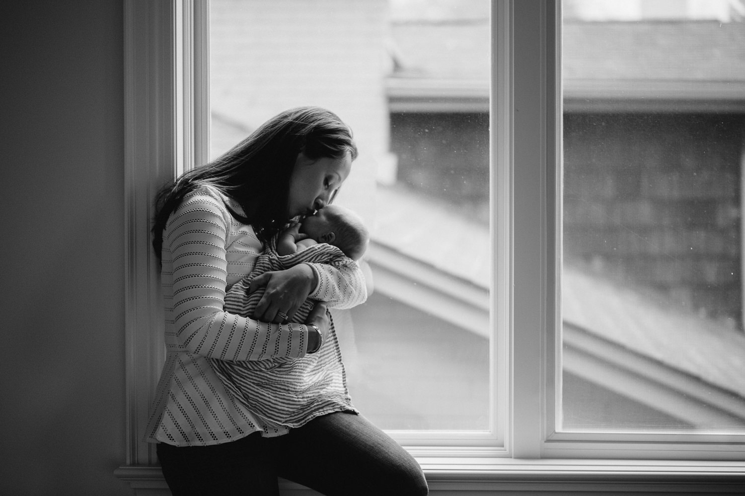 mom kisses 2 week old baby son on forehead, standing on front of window - Stouffville lifestyle photography 