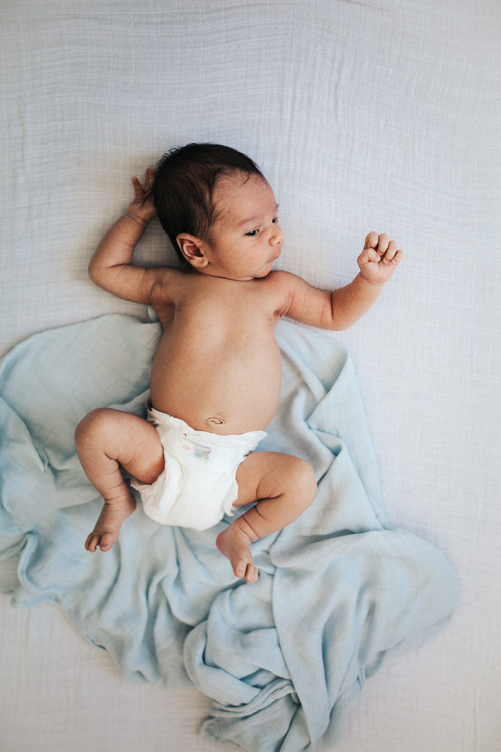 2 week old baby boy in diaper awake, lying on white backdrop with blue swaddle - Barrie Lifestyle Photography 