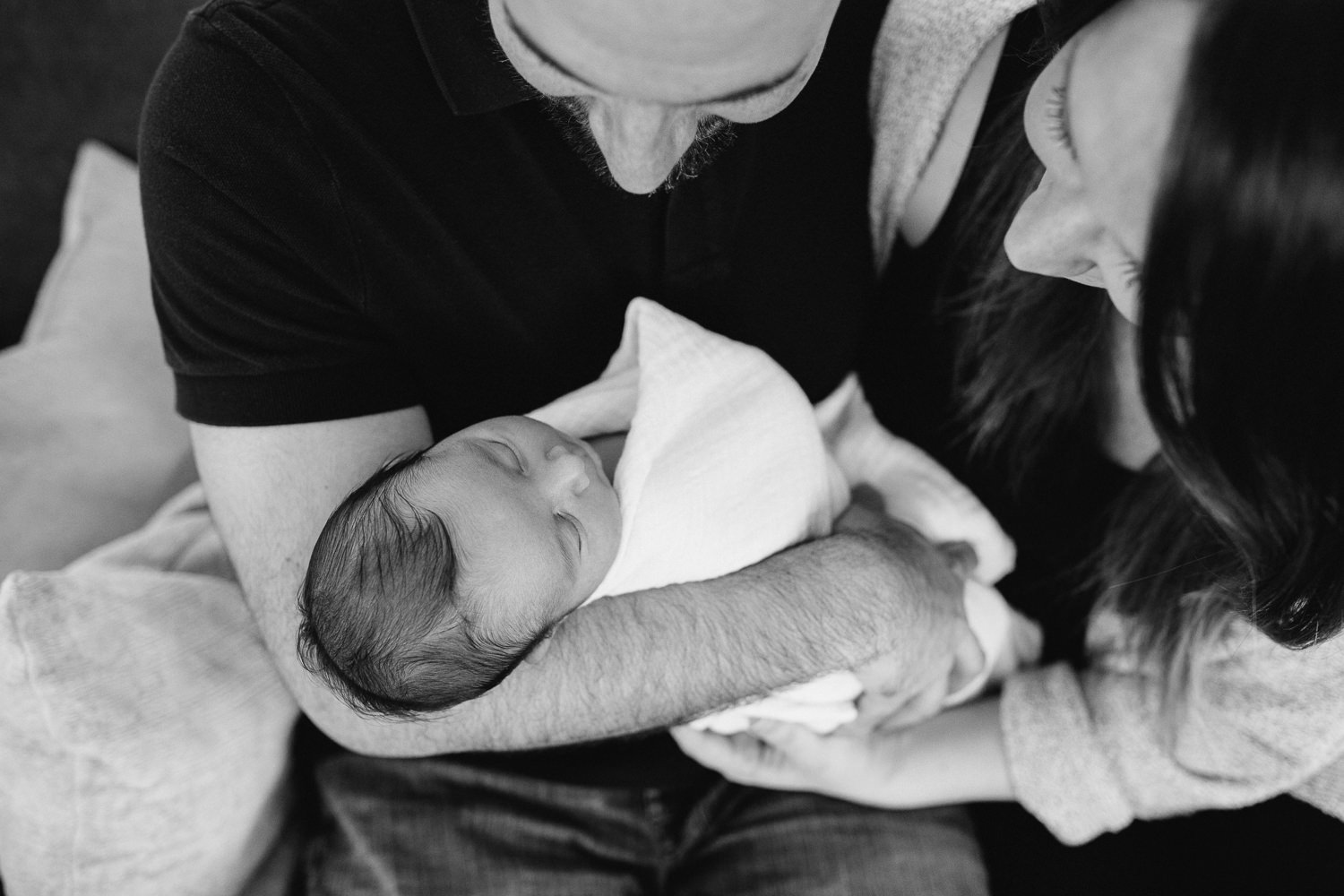 new parents sitting on couch holding and looking at 2 week old baby boy -  Stouffville in-home photos